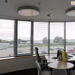 Widok z wnętrza nowoczesnego biura na panoramiczne, łukowe przeszklenie wyposażone w system wewnętrznych żaluzji poziomych C50 Slim. Srebrne, aluminiowe lamele są ustawione w pozycji otwartej, co zapewnia optymalną kontrolę nad dopływem światła dziennego przy zachowaniu widoczności na zewnętrzny parking oraz infrastrukturę miejską. W pomieszczeniu znajduje się stanowisko pracy z biurkiem o jasnym blacie, wyposażone w monitory komputerowe oddzielone żółtym panelem akustycznym oraz ergonomiczne fotele biurowe. Na suficie zamontowano dwie duże, okrągłe oprawy oświetleniowe typu plafon oraz okrągłe anemostaty systemu wentylacji. Estetyka wnętrza jest minimalistyczna, z białymi ścianami i ciemną wykładziną, co podkreśla techniczny charakter zastosowanych osłon okiennych dopasowanych do krzywizny fasady.