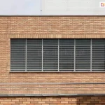 Elewacja nowoczesnego budynku z cegły klinkierowej w kolorze piaskowym, z rzędem dużych okien wyposażonych w zewnętrzne żaluzje fasadowe z poziomymi, aluminiowymi lamelami. Żaluzje zamontowane są na całej długości przeszklenia, z podziałem na segmenty okienne oddzielone pionowymi profilami. Lamele Aluprof ustawione są w pozycji półotwartej, co pozwala na kontrolę dostępu światła do wnętrza. W dolnej części zdjęcia widoczny jest stalowy balkon z balustradą, a po prawej stronie fragment wnęki z lampami elewacyjnymi. Styl budynku sugeruje zastosowanie w obiektach biurowych lub użyteczności publicznej.