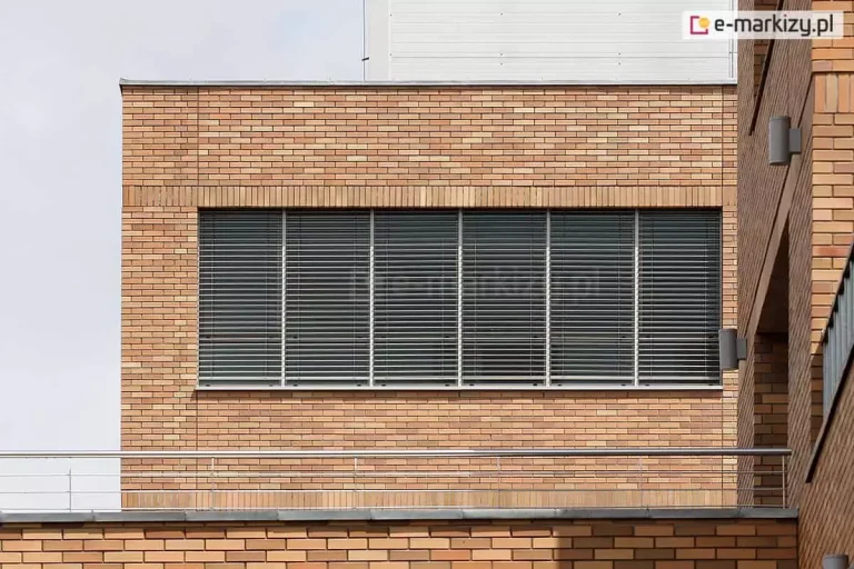 Elewacja nowoczesnego budynku z cegły klinkierowej w kolorze piaskowym, z rzędem dużych okien wyposażonych w zewnętrzne żaluzje fasadowe z poziomymi, aluminiowymi lamelami. Żaluzje zamontowane są na całej długości przeszklenia, z podziałem na segmenty okienne oddzielone pionowymi profilami. Lamele Aluprof ustawione są w pozycji półotwartej, co pozwala na kontrolę dostępu światła do wnętrza. W dolnej części zdjęcia widoczny jest stalowy balkon z balustradą, a po prawej stronie fragment wnęki z lampami elewacyjnymi. Styl budynku sugeruje zastosowanie w obiektach biurowych lub użyteczności publicznej.