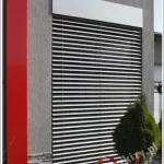 Nowoczesna elewacja budynku z wysokim oknem osłoniętym żaluzją fasadową C50 Selt. System składa się z licznych, poziomych lameli aluminiowych w kolorze srebrnym, które są całkowicie rozwinięte i ustawione w pozycji zamkniętej, co zapewnia pełną ochronę przed słońcem oraz nagrzewaniem wnętrza. Mechanizm żaluzji chowa się pod blachą osłonową w kolorze jasnoszarym, zamontowaną na tle strukturalnego, szarego tynku. Po lewej stronie modułu widoczny jest charakterystyczny, szeroki pas dekoracyjny elewacji w kolorze intensywnej czerwieni, a u podstawy okna znajduje się dekoracyjna podmurówka z kamienia naturalnego oraz nasadzenia roślinne. Detal ten obrazuje estetyczną integrację zewnętrznych żaluzji poziomych z nowoczesną architekturą obiektu, podkreślając ich funkcję termomodernizacyjną i wizualną.