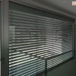 Nowoczesna przestrzeń wewnętrzna z dużym przeszkleniem chronionym przez zewnętrzne żaluzje fasadowe. Aluminiowe lamele żaluzji w ciemnym odcieniu odbijają światło dzienne i zapewniają częściowe zacienienie. Balustrada wykonana z przezroczystego szkła z metalowym uchwytem biegnie wzdłuż korytarza na piętrze. Ściany i sufit w kolorze białym, z wbudowanym oświetleniem punktowym. Po lewej stronie widoczne drzwi z czarną klamką. Przez żaluzje przebija się rozmyty widok na zewnętrzną przestrzeń wokół budynku.