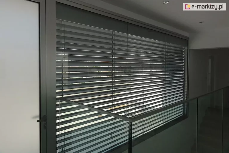 Nowoczesna przestrzeń wewnętrzna z dużym przeszkleniem chronionym przez zewnętrzne żaluzje fasadowe. Aluminiowe lamele żaluzji w ciemnym odcieniu odbijają światło dzienne i zapewniają częściowe zacienienie. Balustrada wykonana z przezroczystego szkła z metalowym uchwytem biegnie wzdłuż korytarza na piętrze. Ściany i sufit w kolorze białym, z wbudowanym oświetleniem punktowym. Po lewej stronie widoczne drzwi z czarną klamką. Przez żaluzje przebija się rozmyty widok na zewnętrzną przestrzeń wokół budynku.