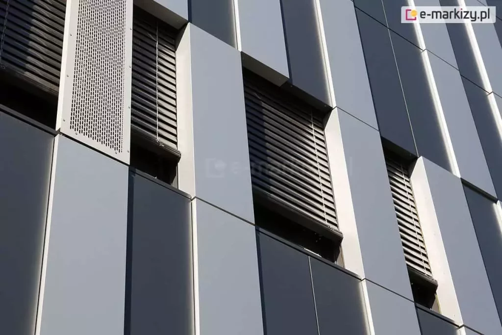 Fragment nowoczesnej elewacji biurowca w ujęciu perspektywicznym od dołu. Fasada składa się z pionowych pasów w dwóch odcieniach szarości oraz wysokich, wąskich wnęk okiennych. W oknach zamontowane są ciemne żaluzje fasadowe z widocznymi linkami prowadzącymi. Po lewej stronie widoczny jest pionowy panel z perforowanej blachy, pełniący funkcję dekoracyjną i wentylacyjną. Gra światła na gładkich płaszczyznach paneli podkreśla geometryczny charakter architektury oraz techniczną precyzję montażu osłon przeciwsłonecznych.