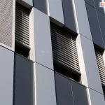 Fragment nowoczesnej elewacji biurowca w ujęciu perspektywicznym od dołu. Fasada składa się z pionowych pasów w dwóch odcieniach szarości oraz wysokich, wąskich wnęk okiennych. W oknach zamontowane są ciemne żaluzje fasadowe z widocznymi linkami prowadzącymi. Po lewej stronie widoczny jest pionowy panel z perforowanej blachy, pełniący funkcję dekoracyjną i wentylacyjną. Gra światła na gładkich płaszczyznach paneli podkreśla geometryczny charakter architektury oraz techniczną precyzję montażu osłon przeciwsłonecznych.