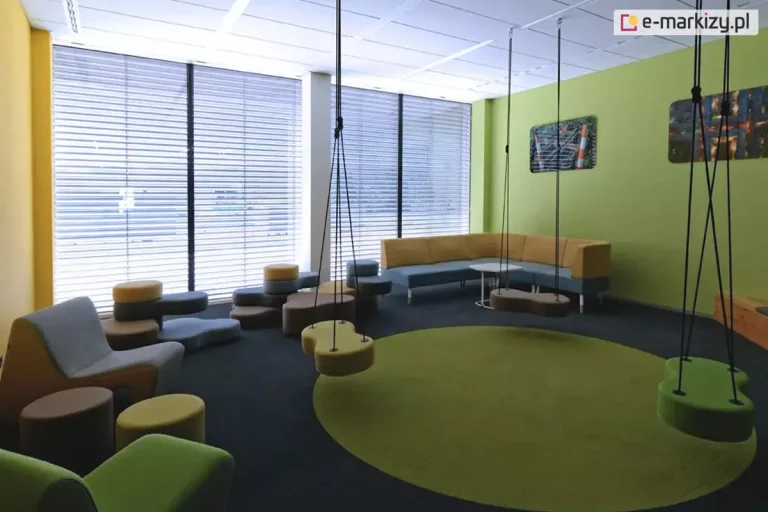 Wnętrze nowoczesnego pokoju wypoczynkowego lub strefy relaksu, z dużymi przeszkleniami od podłogi do sufitu, wyposażonymi w zewnętrzne żaluzje fasadowe z lamelami w kształcie litery C lub Z. Żaluzje zamontowane są w prowadnicach bocznych, zapewniających stabilność i precyzyjne prowadzenie lameli. Lamele wykonane z lakierowanego aluminium, ustawione w pozycji lekko uchylonej, co pozwala na doświetlenie wnętrza przy jednoczesnym ograniczeniu nagrzewania i ochronie przed bezpośrednim światłem słonecznym. Wewnątrz pomieszczenia znajdują się designerskie meble modułowe: tapicerowane fotele, pufy, huśtawki podwieszane do sufitu oraz kanapa narożna, przeznaczone do siedzenia i relaksu. Sufit podwieszany z liniowym oświetleniem LED, ściany pomalowane na kolory żółty i zielono-limonkowy. Żaluzje stanowią element systemu osłon przeciwsłonecznych, poprawiających komfort termiczny i wizualny w budynku.
