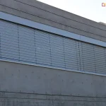 Zewnętrzna żaluzja fasadowa zamontowana w górnej części dużego przeszklenia na elewacji budynku o wykończeniu z betonu architektonicznego. System z aluminiowymi lamelami w kolorze srebrnym, zintegrowany w nowoczesnej wnęce elewacyjnej o prostokątnym kształcie. Lamele ustawione w pozycji półotwartej, umożliwiają częściowy dopływ światła do wnętrza. Widoczne są pionowe taśmy prowadzące i skrzynka maskująca górny mechanizm, dopasowana kolorystycznie do całości systemu. Elewacja z betonu licowego z widocznymi otworami montażowymi i poziomymi podziałami.