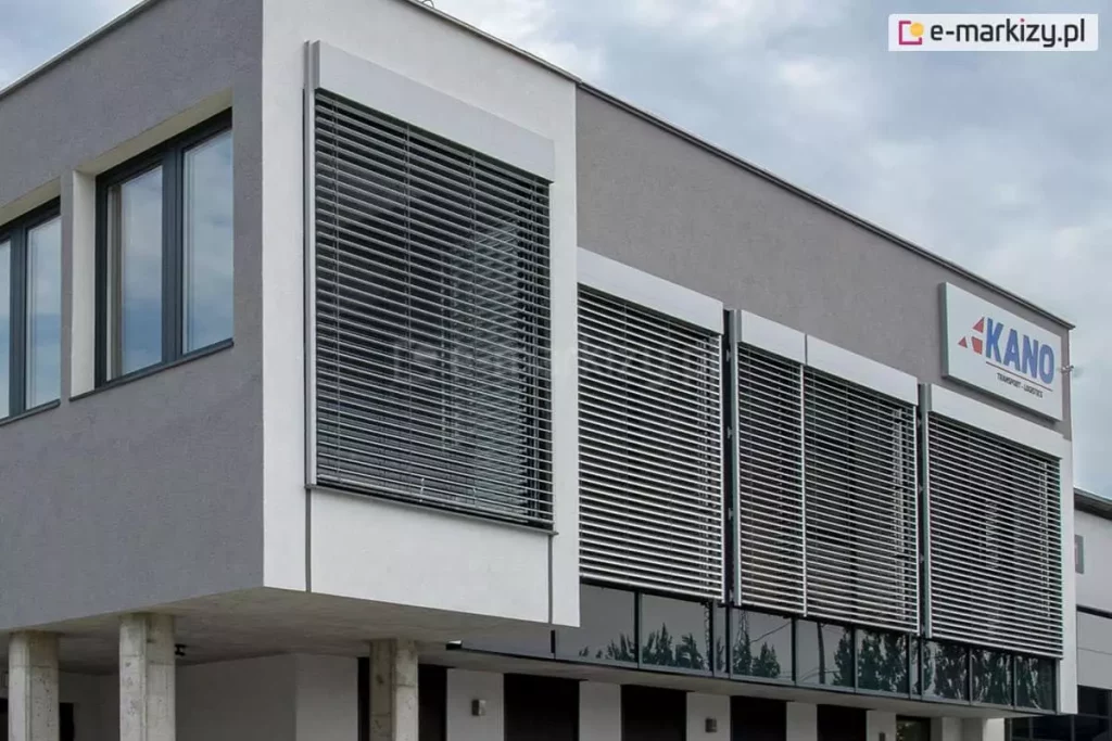 Żaluzja z90 cube z kasetą prostokątną i prowadnicami zewnętrznymi zamontowana na kilku sąsiadujących oknach budynku biurowca o szarej elewacji z widocznym w prawym górnym narożniku logo firmy. Nad budynkiem zachmurzone niego.