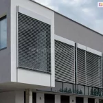 Żaluzja z90 cube z kasetą prostokątną i prowadnicami zewnętrznymi zamontowana na kilku sąsiadujących oknach budynku biurowca o szarej elewacji z widocznym w prawym górnym narożniku logo firmy. Nad budynkiem zachmurzone niego.