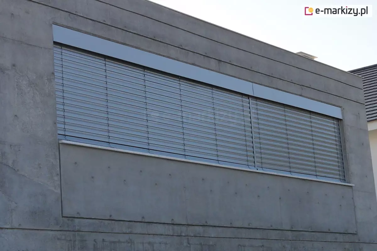 Zewnętrzna żaluzja fasadowa zamontowana w górnej części dużego przeszklenia na elewacji budynku o wykończeniu z betonu architektonicznego. System z aluminiowymi lamelami w kolorze srebrnym, zintegrowany w nowoczesnej wnęce elewacyjnej o prostokątnym kształcie. Lamele ustawione w pozycji półotwartej, umożliwiają częściowy dopływ światła do wnętrza. Widoczne są pionowe taśmy prowadzące i skrzynka maskująca górny mechanizm, dopasowana kolorystycznie do całości systemu. Elewacja z betonu licowego z widocznymi otworami montażowymi i poziomymi podziałami.