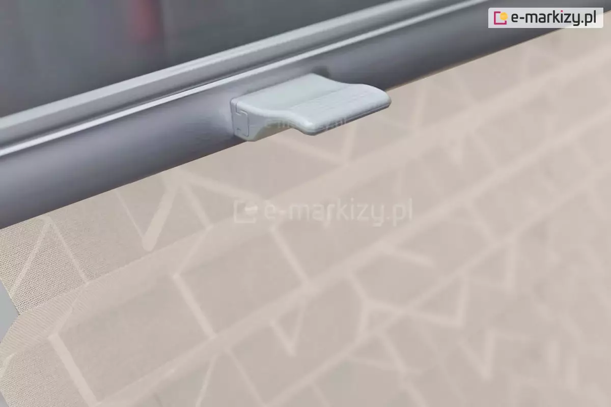 Zdjęcie przedstawia zbliżenie na detal techniczny, jakim jest żaluzja plisowana wyposażona w ergonomiczny uchwyt sterujący. Na pierwszym planie widoczny jest szary profil aluminiowy stanowiący dolną lub górną belkę ruchomą osłony okiennej, do którego przymocowany jest niewielki, jasnoszary element z tworzywa sztucznego służący do ręcznego przesuwania osłony. Materiał, z którego wykonana jest żaluzja plisowana, posiada jasny, beżowy kolor i jest ozdobiony subtelnym, geometrycznym wzorem składającym się z przecinających się linii tworzących kształty trójkątów i rombów. Tkanina wykazuje lekką półprzezroczystość, co jest widoczne dzięki rozproszonemu światłu przechodzącemu przez jej strukturę. W górnej części kadru, powyżej metalowego profilu, znajduje się ciemniejsza, gładka powierzchnia, prawdopodobnie szyba okienna. Całość kompozycji skupia się na funkcjonalności mechanizmu bezsznurkowego, podkreślając nowoczesny design i estetykę wykończenia detali konstrukcyjnych żaluzji.
