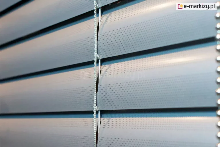 Zbliżenie lameli perforowanych żaluzji aluminiowej z charakterystyczną dziurkowaną strukturą. Zamknięte płatki lameli wpuszczają do pomieszczenia niewielką ilość światła. Pomiędzy lamelami widoczna biała drabinka sznurkowa.