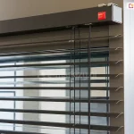 Górna część żaluzji fasadowej C50 Slim Aluprof z widoczną kwadratową kasetą oraz wyprowadzonym z niej systemem sterowania i drabinką sznurkową pomiędzy lamelami w odcieniu grafitu. Kaseta zawieszona na ścianie za pomocą specjalnych uchwytów, które pozostają prawie niewidoczne.