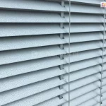 Zdjęcie prezentuje szczegółowe ujęcie z bliska na żaluzje poziome aluminiowe w kolorze jasnoszarym lub srebrnym o unikalnej, teksturowanej powierzchni. Lamele posiadają drobnoziarnisty wzór przypominający nakropienie lub strukturę granitu, co nadaje im matowe wykończenie i nowoczesny wygląd. Żaluzje są ustawione pod skosem w pozycji półotwartej, co pozwala dostrzec ich profil oraz sposób montażu. Przez otwory w lameli przechodzą pionowe elementy konstrukcyjne, czyli drabinka sznurkowa w dopasowanym jasnym kolorze, która utrzymuje listwy w równych odstępach i umożliwia regulację kąta nachylenia. Po prawej stronie widoczny jest pionowy sznurek operacyjny służący do sterowania osłoną. Krawędzie lameli są gładko wykończone i lekko zaokrąglone na rogach. Oświetlenie pada z prawej strony, podkreślając wypukłą strukturę materiału oraz tworząc delikatne cienie pod każdą z listew, co nadaje zdjęciu głębi i uwydatnia techniczną precyzję wykonania produktu.