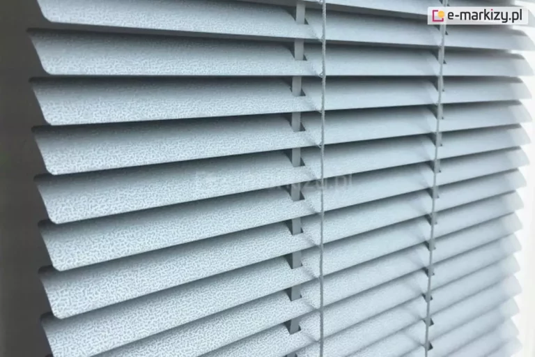 Zdjęcie prezentuje szczegółowe ujęcie z bliska na żaluzje poziome aluminiowe w kolorze jasnoszarym lub srebrnym o unikalnej, teksturowanej powierzchni. Lamele posiadają drobnoziarnisty wzór przypominający nakropienie lub strukturę granitu, co nadaje im matowe wykończenie i nowoczesny wygląd. Żaluzje są ustawione pod skosem w pozycji półotwartej, co pozwala dostrzec ich profil oraz sposób montażu. Przez otwory w lameli przechodzą pionowe elementy konstrukcyjne, czyli drabinka sznurkowa w dopasowanym jasnym kolorze, która utrzymuje listwy w równych odstępach i umożliwia regulację kąta nachylenia. Po prawej stronie widoczny jest pionowy sznurek operacyjny służący do sterowania osłoną. Krawędzie lameli są gładko wykończone i lekko zaokrąglone na rogach. Oświetlenie pada z prawej strony, podkreślając wypukłą strukturę materiału oraz tworząc delikatne cienie pod każdą z listew, co nadaje zdjęciu głębi i uwydatnia techniczną precyzję wykonania produktu.