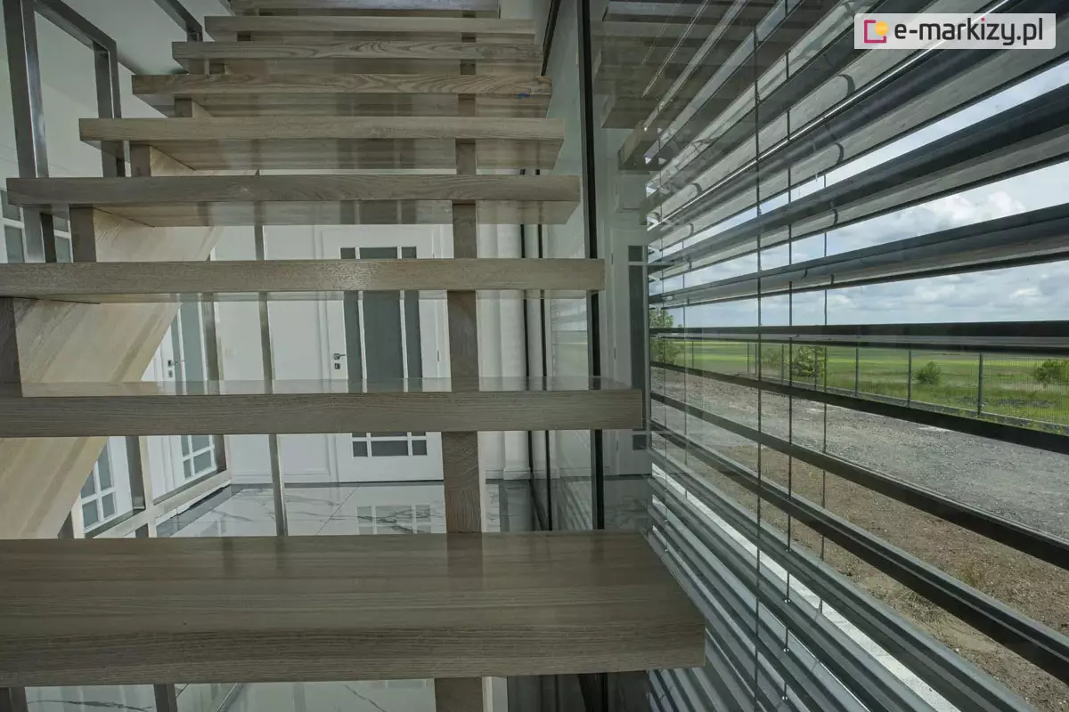 Nowoczesna przestrzeń wewnętrzna z otwartymi schodami drewnianymi typu wspornikowego, umieszczonymi przy przeszklonej ścianie osłoniętej systemem żaluzji fasadowych zewnętrznych. Lamele żaluzji wykonane z aluminium, zamontowane równolegle i lekko uchylone, tworzą estetyczną osłonę przeciwsłoneczną. Schody prowadzą na wyższe piętro i zostały połączone z przeszkloną balustradą. W tle elegancka część wejściowa z białymi drzwiami i połyskującą posadzką z dużych płyt. Za oknem widoczny krajobraz z polami uprawnymi, żwirem oraz pasem zieleni ogrodowej.