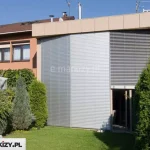Zewnętrzne żaluzje fasadowe aluminiowe Aluprof zamontowane na pełnej wysokości przeszklonej ściany w zabudowie szeregowej, w pozycji całkowicie zasłoniętej z lewej strony oraz częściowo otwartej z prawej strony. Lamele poziome w kolorze jasnoszarym zapewniają skuteczną ochronę przeciwsłoneczną i termiczną. System osłon osadzony na konstrukcji z aluminiowych prowadnic bocznych, wkomponowany w nowoczesną zabudowę elewacyjną z panelami w kolorze piaskowym i dużym przeszkleniem narożnym. Całość otoczona gęstą zielenią ogrodu, z widocznymi tujami, iglakami oraz rozłożonym parasolem tarasowym.