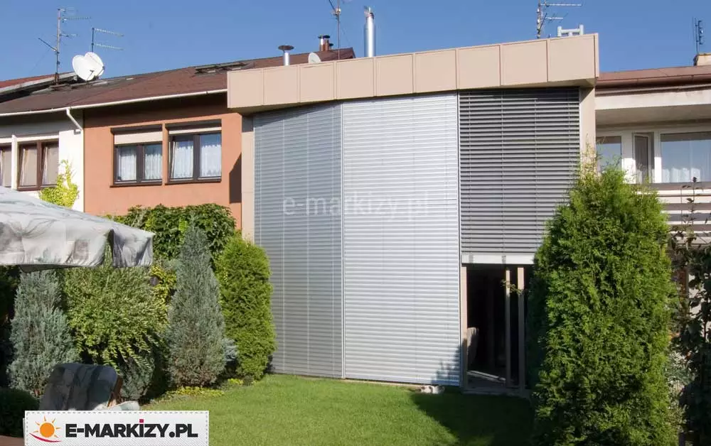 Zewnętrzne żaluzje fasadowe aluminiowe Aluprof zamontowane na pełnej wysokości przeszklonej ściany w zabudowie szeregowej, w pozycji całkowicie zasłoniętej z lewej strony oraz częściowo otwartej z prawej strony. Lamele poziome w kolorze jasnoszarym zapewniają skuteczną ochronę przeciwsłoneczną i termiczną. System osłon osadzony na konstrukcji z aluminiowych prowadnic bocznych, wkomponowany w nowoczesną zabudowę elewacyjną z panelami w kolorze piaskowym i dużym przeszkleniem narożnym. Całość otoczona gęstą zielenią ogrodu, z widocznymi tujami, iglakami oraz rozłożonym parasolem tarasowym.