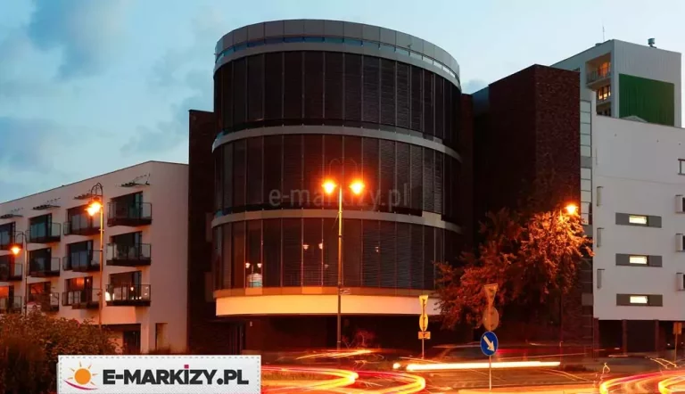 Nowoczesna fasada przeszklonego budynku wielopiętrowego o zaokrąglonym froncie, wyposażona w zewnętrzne żaluzje fasadowe. Żaluzje pokrywają całą powierzchnię szklanych ścian na trzech kondygnacjach, tworząc jednolitą, poziomą strukturę złożoną z równolegle ułożonych lameli aluminiowych w kolorze ciemnoszarym. Lamele są równomiernie rozmieszczone i zamontowane w prowadnicach bocznych, dzięki czemu dopasowują się do krzywizny fasady. Żaluzje są zamontowane we wnękach fasadowych pomiędzy poziomymi, wystającymi pasami konstrukcyjnymi w jasnym kolorze, które dzielą przeszkloną elewację na piętra. W tle widoczna jest część elewacji z ciemnej cegły oraz sąsiadujące budynki mieszkalne – jeden z balkonami, drugi w białym tynku z prostokątnymi oknami. Zdjęcie wykonane o zmierzchu – widać działające oświetlenie uliczne i świetlne smugi przejeżdżających samochodów.