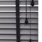 Zdjęcie prezentuje szczegółowe zbliżenie na żaluzje aluminiowe 50 mm w eleganckim, matowym odcieniu szarości lub antracytu. Szerokie, poziome lamele o szerokości pięćdziesięciu milimetrów są ułożone równolegle, tworząc rytmiczną strukturę z wyraźnymi odstępami, przez które prześwituje białe tło. Kluczowym elementem dekoracyjnym i technicznym jest pionowa, szeroka drabinka taśmowa w kolorze głębokiej czerni, wykonana z gęsto tkanej tkaniny o widocznym splocie, która podtrzymuje lamele. Po prawej stronie kadru znajdują się trzy czarne sznurki operacyjne o różnej długości, z których każdy zakończony jest ergonomicznym, dekoracyjnym obciążnikiem w kształcie łezki wykonanym z czarnego tworzywa. Sznurki te służą do sterowania mechanizmem podnoszenia oraz kątem nachylenia listew. Kompozycja podkreśla nowoczesny design oraz kontrastowe połączenie kolorystyczne między szarymi lamelami a czarnym osprzętem, co jest charakterystyczne dla systemów, w których wykorzystywana jest żaluzja aluminiowa 50 mm.