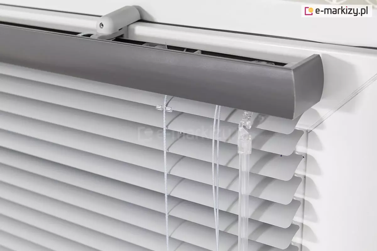 Zbliżenie bezinwazyjnego mocowania żaluzji aluminiowej do skrzydła okna. Żaluzja z aluminiowymi lamelami 25 mm oraz czarną dodatkowa maskownicą drewnianą zawieszona została na skrzydle okna za pomocą białych uchwytów bezinwazyjnych. Po prawej stronie widoczny także system sterowania żaluzją - sznurki oraz pokrętło.
