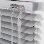 Zbliżenie zakończenia bocznego rynny górnej oraz mechanizmu sterowania żaluzją aluminiową 25 mm, z jasnoszarymi matowymi lamelami, białym sznurkiem oraz lexanowym pokrętłem. Żaluzja zamocowana na białej ramie okna.