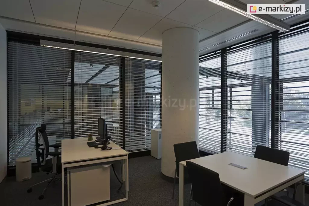 Widok na nowoczesną przestrzeń biurową typu open space, w której zastosowano system żaluzji fasadowych C50 Slim na wielkoformatowych przeszkleniach. Aluminiowe lamele w kolorze srebrnym są ustawione w pozycji otwartej, co pozwala na optymalne doświetlenie wnętrza światłem dziennym przy jednoczesnym ograniczeniu uciążliwych odblasków na ekranach monitorów. Pomieszczenie charakteryzuje się surowym, industrialnym designem z centralnie umieszczonym betonowym słupem konstrukcyjnym, ciemną wykładziną oraz nowoczesnym oświetleniem liniowym zamontowanym pod kasetonowym sufitem. Wyposażenie obejmuje minimalistyczne białe biurka, ergonomiczne czarne fotele biurowe oraz systemowe szafki na dokumenty. Precyzyjna regulacja kąta nachylenia lameli zapewnia komfort pracy oraz prywatność, harmonijnie współgrając z technicznym charakterem architektury budynku.