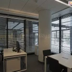 Widok na nowoczesną przestrzeń biurową typu open space, w której zastosowano system żaluzji fasadowych C50 Slim na wielkoformatowych przeszkleniach. Aluminiowe lamele w kolorze srebrnym są ustawione w pozycji otwartej, co pozwala na optymalne doświetlenie wnętrza światłem dziennym przy jednoczesnym ograniczeniu uciążliwych odblasków na ekranach monitorów. Pomieszczenie charakteryzuje się surowym, industrialnym designem z centralnie umieszczonym betonowym słupem konstrukcyjnym, ciemną wykładziną oraz nowoczesnym oświetleniem liniowym zamontowanym pod kasetonowym sufitem. Wyposażenie obejmuje minimalistyczne białe biurka, ergonomiczne czarne fotele biurowe oraz systemowe szafki na dokumenty. Precyzyjna regulacja kąta nachylenia lameli zapewnia komfort pracy oraz prywatność, harmonijnie współgrając z technicznym charakterem architektury budynku.