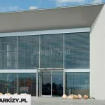 Nowoczesny budynek o minimalistycznej bryle, z prostą elewacją w białym kolorze i dużym, dwukondygnacyjnym przeszkleniem na froncie. Przeszklona fasada została wyposażona w zewnętrzne żaluzje fasadowe z poziomymi lamelami aluminiowymi w kolorze srebrnoszarym. Lamele są równoległe, gęsto rozmieszczone i osadzone w bocznych prowadnicach, zasłaniają całą przeszkloną powierzchnię zarówno na parterze, jak i na piętrze. Wejście do budynku znajduje się centralnie – składa się z dwuskrzydłowych, szklanych drzwi automatycznych, nad którymi zamontowano daszek ze szkła wsparty na stalowych wspornikach. W dolnej części elewacji widoczne są ozdobne, jasne kamienie ułożone na podłożu żwirowym. Dach budynku jest płaski, z charakterystycznym wysunięciem tworzącym głęboki, cieniujący okap. W tle widoczne są inne obiekty przemysłowe i zabudowa jednorodzinna.