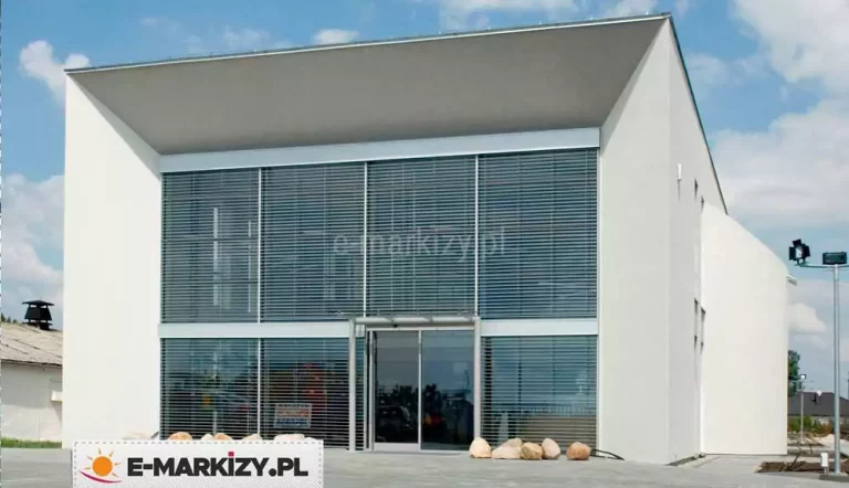 Nowoczesny budynek o minimalistycznej bryle, z prostą elewacją w białym kolorze i dużym, dwukondygnacyjnym przeszkleniem na froncie. Przeszklona fasada została wyposażona w zewnętrzne żaluzje fasadowe z poziomymi lamelami aluminiowymi w kolorze srebrnoszarym. Lamele są równoległe, gęsto rozmieszczone i osadzone w bocznych prowadnicach, zasłaniają całą przeszkloną powierzchnię zarówno na parterze, jak i na piętrze. Wejście do budynku znajduje się centralnie – składa się z dwuskrzydłowych, szklanych drzwi automatycznych, nad którymi zamontowano daszek ze szkła wsparty na stalowych wspornikach. W dolnej części elewacji widoczne są ozdobne, jasne kamienie ułożone na podłożu żwirowym. Dach budynku jest płaski, z charakterystycznym wysunięciem tworzącym głęboki, cieniujący okap. W tle widoczne są inne obiekty przemysłowe i zabudowa jednorodzinna.