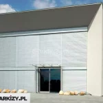 Nowoczesny, minimalistyczny budynek o białej elewacji z płaskim, jednospadowym dachem i dużą przeszkloną fasadą frontową. Na całej wysokości elewacji frontowej zamontowane są zewnętrzne żaluzje fasadowe C80 Cube z widocznymi kasetami. Składają się one z poziomych, wąskich aluminiowych lamel w kolorze srebrnym, prowadnic listwowych modułowych oraz zabudowanych skrzynek osłonowych – całkowicie zlicowanych z elewacją, co podkreśla estetykę i czystość formy. Żaluzje pokrywają dwa duże, pionowe segmenty przeszkleń – po lewej i prawej stronie fasady – rozdzielone pionowym słupem konstrukcyjnym. W centralnej części widoczne jest główne wejście z podwójnymi szklanymi drzwiami, nad którymi znajduje się szklany daszek wsparty stalowymi słupkami. Przed budynkiem ułożono nieregularne głazy dekoracyjne wzdłuż elewacji. Tło stanowi niebieskie, lekko zachmurzone niebo, a całość otacza utwardzony plac.