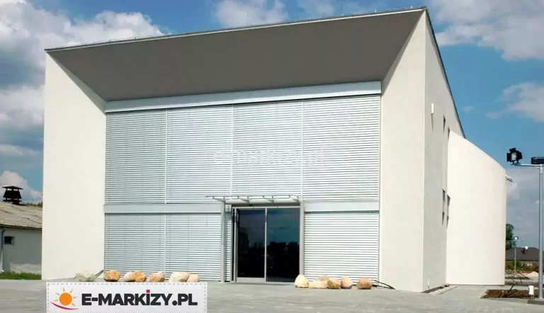 Nowoczesny, minimalistyczny budynek o białej elewacji z płaskim, jednospadowym dachem i dużą przeszkloną fasadą frontową. Na całej wysokości elewacji frontowej zamontowane są zewnętrzne żaluzje fasadowe C80 Cube z widocznymi kasetami. Składają się one z poziomych, wąskich aluminiowych lamel w kolorze srebrnym, prowadnic listwowych modułowych oraz zabudowanych skrzynek osłonowych – całkowicie zlicowanych z elewacją, co podkreśla estetykę i czystość formy. Żaluzje pokrywają dwa duże, pionowe segmenty przeszkleń – po lewej i prawej stronie fasady – rozdzielone pionowym słupem konstrukcyjnym. W centralnej części widoczne jest główne wejście z podwójnymi szklanymi drzwiami, nad którymi znajduje się szklany daszek wsparty stalowymi słupkami. Przed budynkiem ułożono nieregularne głazy dekoracyjne wzdłuż elewacji. Tło stanowi niebieskie, lekko zachmurzone niebo, a całość otacza utwardzony plac.