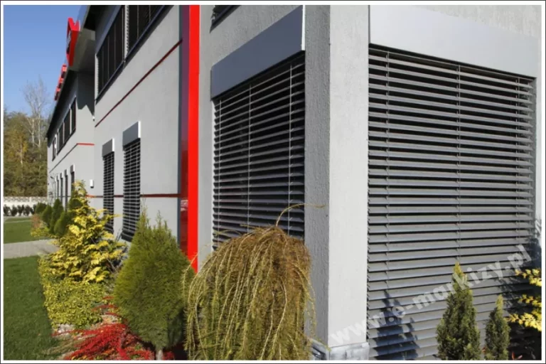 Widok na elewację nowoczesnego budynku biurowego lub usługowego, na której zainstalowano system żaluzji fasadowych C50L. Ciemne, antracytowe lamele są zamontowane w dedykowanych kasetach osłonowych, które licują się z płaszczyzną jasnoszarej, tynkowanej ściany. Żaluzje w pozycji zamkniętej całkowicie przysłaniają wysokie przeszklenia, zapewniając maksymalną ochronę przed nagrzewaniem wnętrza oraz gwarantując prywatność. Architektura obiektu wyróżnia się nowoczesnymi detalami, takimi jak pionowe czerwone pasy dekoracyjne oraz systemowe obróbki blacharskie. Wokół budynku znajduje się zadbana zieleń niska – ozdobne iglaki, krzewy oraz trawnik, co podkreśla estetyczne walory integracji zewnętrznych osłon przeciwsłonecznych z otoczeniem biznesowym. Perspektywa zdjęcia ukazuje powtarzalność modułów żaluzji na całej długości fasady, co dokumentuje kompleksową realizację systemu zaciemnienia dla dużego obiektu.