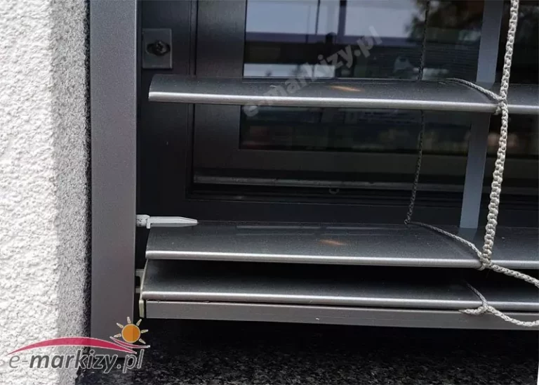 Zbliżenie na dolną część zewnętrznej żaluzji fasadowej C80, zamontowanej na elewacji budynku. Widoczne są poziome aluminiowe lamele o delikatnym łukowym wyprofilowaniu w kształcie litery „C”, które umożliwiają regulację kąta nachylenia w celu kontrolowania ilości światła wpadającego do wnętrza. Lamele są w kolorze antracytowym (zbliżonym do RAL 7016) z lekko połyskującym, lakierowanym wykończeniem. Na powierzchni lameli widoczne są drobne refleksy świetlne oraz naturalne załamania odbicia otoczenia. Po prawej stronie zdjęcia znajduje się system sznurkowy żaluzji — liny prowadzące oraz drabinki tekstylne (taśmy) wykonane z odpornego na warunki atmosferyczne materiału syntetycznego w kolorze jasnoszarym. Sznury są estetycznie napięte i przymocowane do lameli, zapewniając ich równomierne unoszenie i opuszczanie. Po lewej stronie widoczna jest prowadnica boczna wykonana z ekstrudowanego aluminium, montowana we wnęce okiennej. Jej powierzchnia jest gładka, lakierowana proszkowo na kolor grafitowy, pasujący kolorystycznie do ramy okna i żaluzji. Wewnątrz prowadnicy zamontowany jest plastikowy element dystansujący (prowadnik) utrzymujący odpowiednie położenie lameli w trakcie ruchu. Dolna listwa końcowa wykonana z profilu aluminiowego stabilizuje całą konstrukcję. W tle widać szybę okna z odbiciem zewnętrznego otoczenia oraz częściowo widoczne wnętrze pomieszczenia. Całość zamontowana na elewacji z tynkiem strukturalnym w kolorze jasnoszarym oraz z parapetem wykonanym z ciemnego kamienia naturalnego lub konglomeratu. Zdjęcie przedstawia wysoką jakość wykonania i precyzyjne spasowanie elementów systemu osłonowego.