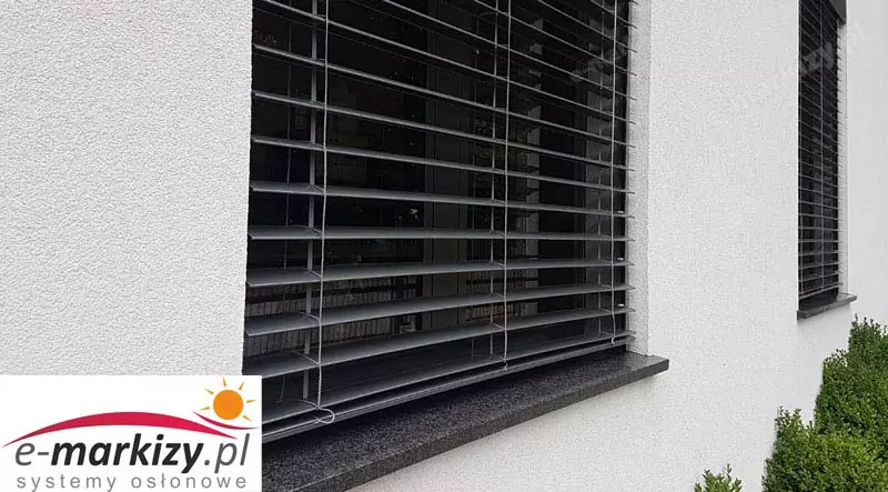 Zbliżenie na zewnętrzne żaluzje fasadowe zamontowane na oknach elewacji budynku mieszkalnego. System składa się z poziomych aluminiowych lameli typu C, ustawionych w pozycji lekko uchylonej, co umożliwia precyzyjne regulowanie dostępu światła dziennego. Lamele utrzymywane są przez system taśm tekstylnych (drabinki) oraz prowadzone bocznie przy pomocy linek stalowych z tworzywową powłoką ochronną. Konstrukcja żaluzji osadzona na elewacji o tynku strukturalnym w kolorze białym, z parapetem wykonanym z polerowanego kamienia granitowego w odcieniu ciemnoszarym. W prawym dolnym rogu widoczna część przydomowego ogrodu z przystrzyżonymi krzewami. Żaluzje spełniają funkcję osłony przeciwsłonecznej i termicznej, jednocześnie podkreślając nowoczesny charakter architektury budynku.