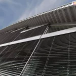 Zbliżenie na fragment nowoczesnej fasady budynku pod kątem, prezentujące instalację żaluzji fasadowych pod zadaszeniem o profilu trapezowym. Lamele w kolorze ciemnoantracytowym są całkowicie opuszczone i uchylone tworząc barierę przeciwsłoneczną na dużych przeszkleniach. Widoczne są srebrne kasety górne o charakterystycznym półokrągłym kształcie (OVAL) oraz system prowadnic bocznych stabilizujących konstrukcję. Nad żaluzjami znajdują się trójkątne doświetla okienne dostosowane do skosu dachu. Ujęcie techniczne podkreśla precyzyjne dopasowanie modułów żaluzji do podziałów okiennych oraz sposób wykończenia krawędzi dachu metalowym opierzeniem.