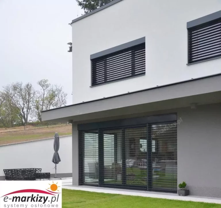 Nowoczesny budynek mieszkalny w stylu minimalistycznym, z zainstalowanymi zewnętrznymi żaluzjami fasadowymi C80 w kolorze antracytowym. Na zdjęciu widać dwie kondygnacje: na piętrze zamontowano dwie mniejsze żaluzje nadokienne, a na parterze jedną dużą, obejmującą całą powierzchnię drzwi tarasowych typu HS. Lamele żaluzji są wykonane z wyprofilowanego aluminium, o kształcie litery "C", co umożliwia płynną regulację kąta ich nachylenia, a tym samym kontrolę natężenia światła wpadającego do wnętrza budynku. Lamele są lakierowane proszkowo na kolor ciemnoszary (RAL 7016), z lekkim połyskiem. Widoczna jest dolna listwa końcowa aluminiowa, która pełni funkcję stabilizującą i obciążającą system. Każda z żaluzji posiada górną osłonę (blacha osłonowa), wykonaną z metalu, również w kolorze antracytowym, ukrywającą mechanizm podnoszenia i opuszczania lameli. Lamele prowadzone są wzdłuż systemu prowadnic bocznych, co zapewnia ich stabilność, nawet przy większym wietrze. System najprawdopodobniej obsługiwany jest elektrycznie – nie są widoczne żadne ręczne mechanizmy sterujące. Budynek ma elewację w dwóch odcieniach – wyższe partie są wykończone w kolorze białym, natomiast dolne fragmenty, obejmujące taras i wnękę okienną, w szarym. Całość utrzymana jest w estetyce nowoczesnej architektury jednorodzinnej, z dużymi przeszkleniami. Przed domem znajduje się zadbany trawnik, a w tle widoczna jest minimalistyczna strefa wypoczynkowa z meblami ogrodowymi i zamkniętym parasolem.