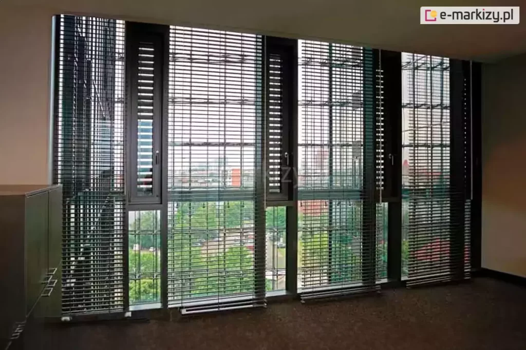 Widok wnętrza nowoczesnego biurowca na ścianę z wysokim przeszkleniem, osłoniętą przez system wielomodułowych żaluzji fasadowych C50L. Aluminiowe lamele w kolorze ciemnego grafitu są ustawione w pozycji otwartej, co pozwala na przenikanie rozproszonego światła dziennego i oferuje panoramiczny widok na miejską zieleń oraz sąsiednią zabudowę. Żaluzje są zintegrowane z pionowymi profilami okiennymi, a każda sekcja posiada indywidualny system prowadzenia linkowego, zapewniający stabilność osłon. Wnętrze charakteryzuje się biurową stylistyką z ciemną wykładziną podłogową i minimalistycznymi meblami, co w połączeniu z precyzyjną geometrią żaluzji podkreśla techniczny i profesjonalny charakter przestrzeni. System ten skutecznie wspiera zarządzanie energią cieplną budynku poprzez redukcję nadmiernego nasłonecznienia dużych powierzchni szklanych.