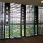 Widok wnętrza nowoczesnego biurowca na ścianę z wysokim przeszkleniem, osłoniętą przez system wielomodułowych żaluzji fasadowych C50L. Aluminiowe lamele w kolorze ciemnego grafitu są ustawione w pozycji otwartej, co pozwala na przenikanie rozproszonego światła dziennego i oferuje panoramiczny widok na miejską zieleń oraz sąsiednią zabudowę. Żaluzje są zintegrowane z pionowymi profilami okiennymi, a każda sekcja posiada indywidualny system prowadzenia linkowego, zapewniający stabilność osłon. Wnętrze charakteryzuje się biurową stylistyką z ciemną wykładziną podłogową i minimalistycznymi meblami, co w połączeniu z precyzyjną geometrią żaluzji podkreśla techniczny i profesjonalny charakter przestrzeni. System ten skutecznie wspiera zarządzanie energią cieplną budynku poprzez redukcję nadmiernego nasłonecznienia dużych powierzchni szklanych.