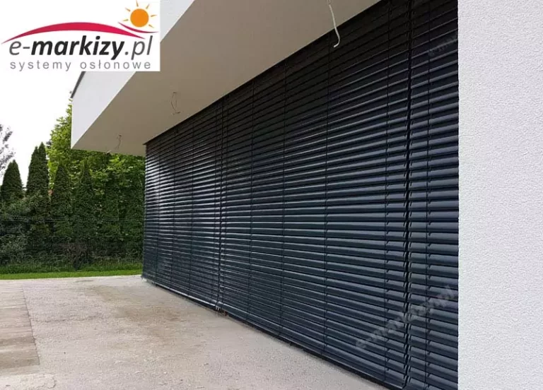 Zewnętrzne żaluzje fasadowe Z90 Selt w pozycji zamkniętej, zamontowane na dużej powierzchni szklanej w nowoczesnym budynku mieszkalnym. Konstrukcja obejmuje jedno długie okno tarasowe, które zostało w całości osłonięte systemem lamelowym wykonanym z aluminium o profilu w kształcie litery „Z”. Lamele są ustawione poziomo, ciasno do siebie przylegają, co zapewnia maksymalne zaciemnienie wnętrza oraz podwyższoną izolację termiczną. Lamele posiadają boczne prowadzenie za pomocą stalowych linek w powłoce ochronnej, które odpowiadają za stabilność osłony podczas wiatru oraz precyzyjne prowadzenie w trakcie podnoszenia i opuszczania. W górnej części fasady widoczna zabudowa podtynkowa – skrzynka rewizyjna została całkowicie ukryta w ociepleniu elewacji, co podnosi estetykę i pozwala na integrację systemu z nowoczesnym designem budynku. Elewacja budynku wykonana jest z białego tynku strukturalnego, co kontrastuje z antracytowym kolorem lamel (prawdopodobnie RAL 7016). Na pierwszym planie znajduje się przestronny taras wykonany z betonu, a w tle – zieleń ogrodu i żywopłot. Widoczne w stropie przewody prawdopodobnie będą służyły do zainstalowania zewnętrznego oświetlenia.