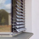 Zbliżenie na zewnętrzną żaluzję fasadową zamontowaną na oknie w elewacji budynku. Przedstawiony system składa się z aluminiowych lameli w kształcie litery C – lekko wygiętych, o delikatnym profilu umożliwiającym precyzyjną regulację dopływu światła. Lamele ustawione są w pozycji półotwartej, co pozwala na kontrolowany dostęp światła dziennego przy zachowaniu prywatności we wnętrzu. Każda lamela zamocowana jest w tekstylnej drabince, prowadzonej pionowo po obu stronach, wykonanej z wytrzymałych materiałów odpornych na warunki atmosferyczne. Dodatkowo, po bokach żaluzji zastosowano stalowe linki napinające (system prowadzenia bocznego), które stabilizują całą konstrukcję i zapobiegają jej kołysaniu pod wpływem wiatru. Dolna szyna obciążająca wykonana z aluminium widoczna jest w dolnej części systemu – zakończona dwoma punktami mocowania do ściany. Okno posiada nowoczesną, aluminiową ramę z szybą refleksyjną, natomiast elewacja wykończona jest jasnym tynkiem strukturalnym. Rozwiązanie to zapewnia funkcjonalną ochronę przeciwsłoneczną, poprawia efektywność energetyczną budynku oraz nadaje mu nowoczesny, techniczny charakter. Szczególnie polecane w budynkach energooszczędnych i pasywnych.