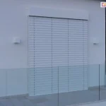 Nowoczesny taras z białą elewacją budynku, na której zamontowana jest Żaluzja fasadowa Z90 w kolorze białym. Żaluzja jest całkowicie opuszczona, zasłaniając duże przeszklenie tarasowe. Powierzchnia tarasu wyłożona jest jasnoszarymi, wielkoformatowymi płytami gresowymi. Po prawej stronie zdjęcia, przy szklanej balustradzie, stoi biała, wysoka, pusta donica. Po lewej stronie, za narożnikiem ściany stoi wysoka czarna donica z drzewkiem ozdobnym. Na elewacji powyżej rolety oraz obok niej znajdują się białe, kwadratowe oprawy oświetlenia zewnętrznego. Taras otacza minimalistyczna szklana balustrada, a w tle za nim widoczne sa drzewa i niebo.