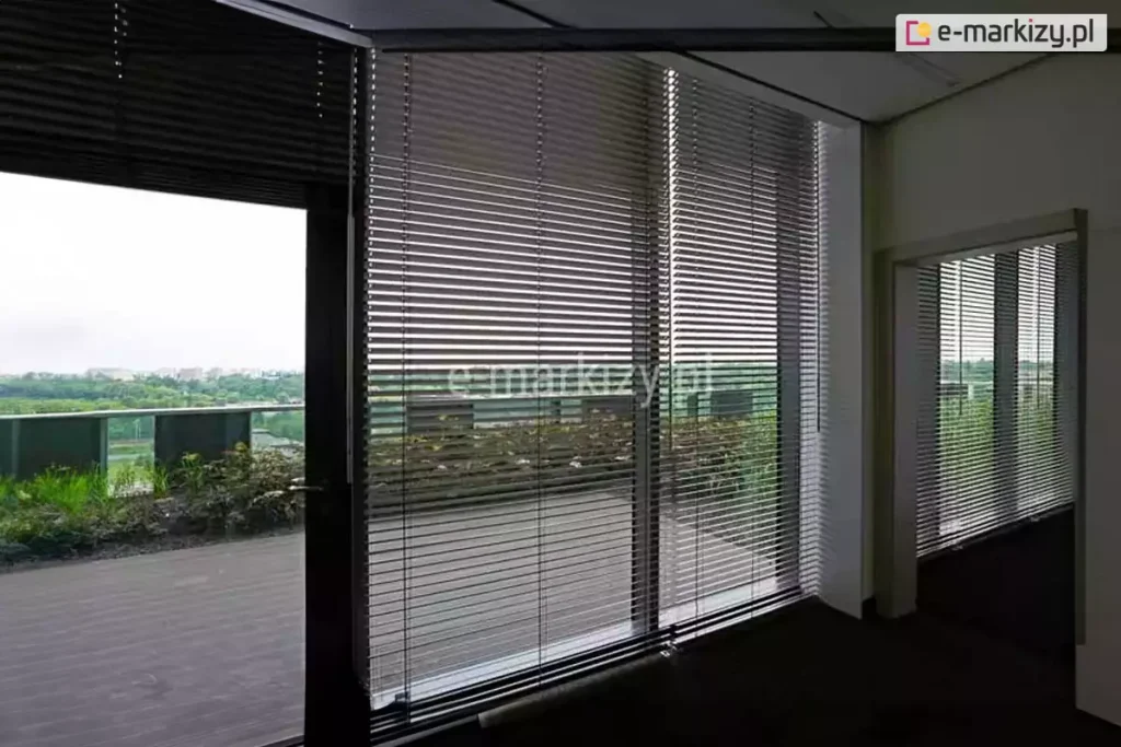 Widok z wnętrza nowoczesnego biura na system wewnętrznych żaluzji fasadowych C50 Aluprof zamontowanych na wielkoformatowych przeszkleniach tarasowych. Srebrne, aluminiowe lamele o wysokim połysku są ustawione w pozycji otwartej, co pozwala na precyzyjne filtrowanie światła dziennego i redukcję nagrzewania pomieszczenia przy zachowaniu widoczności na zewnętrzny taras widokowy. Przez żaluzje widoczna jest balustrada tarasu, niska zieleń ozdobna oraz rozległa panorama miasta w tle. Konstrukcja osłon obejmuje system prowadzenia bocznego, który stabilizuje żaluzje przy otwieraniu drzwi balkonowych. Ciemna podłoga biura kontrastuje z jasnym wykończeniem sufitu i ścian, podkreślając techniczny, a zarazem elegancki charakter zastosowanego systemu zaciemnienia fasadowego.