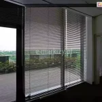 Widok z wnętrza nowoczesnego biura na system wewnętrznych żaluzji fasadowych C50 Aluprof zamontowanych na wielkoformatowych przeszkleniach tarasowych. Srebrne, aluminiowe lamele o wysokim połysku są ustawione w pozycji otwartej, co pozwala na precyzyjne filtrowanie światła dziennego i redukcję nagrzewania pomieszczenia przy zachowaniu widoczności na zewnętrzny taras widokowy. Przez żaluzje widoczna jest balustrada tarasu, niska zieleń ozdobna oraz rozległa panorama miasta w tle. Konstrukcja osłon obejmuje system prowadzenia bocznego, który stabilizuje żaluzje przy otwieraniu drzwi balkonowych. Ciemna podłoga biura kontrastuje z jasnym wykończeniem sufitu i ścian, podkreślając techniczny, a zarazem elegancki charakter zastosowanego systemu zaciemnienia fasadowego.