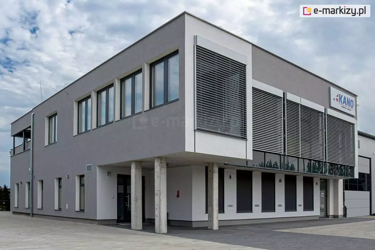 Duży piętrowy budynek, siedziba firmy, której logo umieszczono na fasadzie w szarym kolorze. Okna na piętrze po prawej stronie zostały osłonięte przed słońcem żaluzjami fasadowymi Z90 Cube z prowadnicami na zewnątrz dopasowanymi kolorystycznie do barw elewacji. Nad budynkiem widoczne zachmurzone niebo.