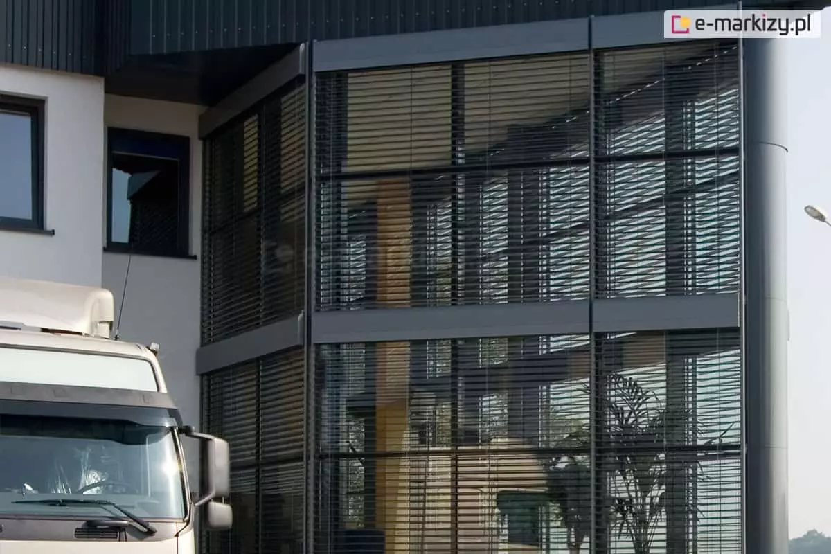 Nowoczesna fasada narożna budynku biurowo-przemysłowego z dużymi, przeszklonymi powierzchniami okiennymi. Okna wyposażone są w zewnętrzne żaluzje fasadowe C80 Oval, wykonane z poziomych aluminiowych lameli w kolorze grafitowym. Lamele są częściowo uchylone, umożliwiając dopływ światła dziennego przy jednoczesnym ograniczeniu nagrzewania wnętrza. Konstrukcja żaluzji zamocowana jest na systemie prowadnic pionowych zintegrowanych z fasadą. Na zdjęciu widoczna jest również bryła budynku z ciemnoszarymi panelami elewacyjnymi oraz białymi ścianami z klasycznymi oknami. Wewnątrz, przez szybę, dostrzec można drewniane słupy konstrukcyjne oraz roślinę doniczkową. Na pierwszym planie, w dolnym lewym rogu, częściowo widoczna jest biała ciężarówka zaparkowana przed budynkiem.