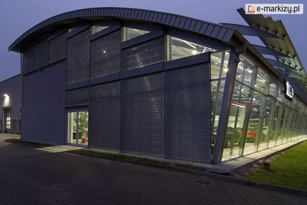 Nowoczesny, dwukondygnacyjny salon samochodowy o przeszklonej elewacji z zainstalowanymi zewnętrznymi żaluzjami fasadowymi C80 Oval w kolorze ciemnoszarym (antracytowym). Zdjęcie wykonane o zmierzchu ukazuje kontrast między oświetlonym wnętrzem salonu a zasłoniętą częścią elewacji. Żaluzje fasadowe o poziomych lamelach pokrywają znaczną część frontowej ściany budynku, skutecznie przesłaniając wysokie przeszklenia. Budynek charakteryzuje się półokrągłym, łukowym dachem z metalowym wykończeniem oraz widoczną stalową konstrukcją wsporczą. Prawa strona salonu pozostaje w pełni przeszklona i nachylona pod kątem, eksponując zaparkowane wewnątrz samochody osobowe (widoczne modele w kolorach czerwonym i ciemnym). Wejście do obiektu stanowią przeszklone drzwi, obok których widać fragment aranżacji wnętrza z rośliną doniczkową. Przed budynkiem znajduje się nawierzchnia z kostki brukowej oraz pas zieleni z trawnikiem. System osłon przeciwsłonecznych jest zintegrowany z architekturą obiektu, podkreślając jego industrialny i profesjonalny charakter.