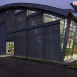 Nowoczesny, dwukondygnacyjny salon samochodowy o przeszklonej elewacji z zainstalowanymi zewnętrznymi żaluzjami fasadowymi C80 Oval w kolorze ciemnoszarym (antracytowym). Zdjęcie wykonane o zmierzchu ukazuje kontrast między oświetlonym wnętrzem salonu a zasłoniętą częścią elewacji. Żaluzje fasadowe o poziomych lamelach pokrywają znaczną część frontowej ściany budynku, skutecznie przesłaniając wysokie przeszklenia. Budynek charakteryzuje się półokrągłym, łukowym dachem z metalowym wykończeniem oraz widoczną stalową konstrukcją wsporczą. Prawa strona salonu pozostaje w pełni przeszklona i nachylona pod kątem, eksponując zaparkowane wewnątrz samochody osobowe (widoczne modele w kolorach czerwonym i ciemnym). Wejście do obiektu stanowią przeszklone drzwi, obok których widać fragment aranżacji wnętrza z rośliną doniczkową. Przed budynkiem znajduje się nawierzchnia z kostki brukowej oraz pas zieleni z trawnikiem. System osłon przeciwsłonecznych jest zintegrowany z architekturą obiektu, podkreślając jego industrialny i profesjonalny charakter.