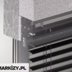 Przekrój montażowy górnej części żaluzji fasadowej zewnętrznej, ukazujący detale techniczne osadzenia systemu w elewacji budynku. Widoczna jest warstwa elewacyjna z tynku strukturalnego oraz fragment warstwy izolacyjnej pod spodem. Wnęka nadprożowa została przygotowana pod montaż osłony – żaluzja Aluprof jest zamocowana w skrzynce aluminiowej, częściowo ukrytej w warstwie ocieplenia. Wewnątrz skrzynki znajduje się górny mechanizm żaluzji, z widoczną listwą montażową, przewodem elektrycznym zasilającym napęd (silnik) oraz elementami mocującymi. Żaluzja składa się z poziomych lameli aluminiowych typu C, w kolorze ciemnoszarym, zawieszonych na taśmach tekstylnych. Część konstrukcyjna oraz prowadnica boczna zostały pokazane w przekroju transparentnym, aby ukazać sposób montażu i przebieg mechanizmu.