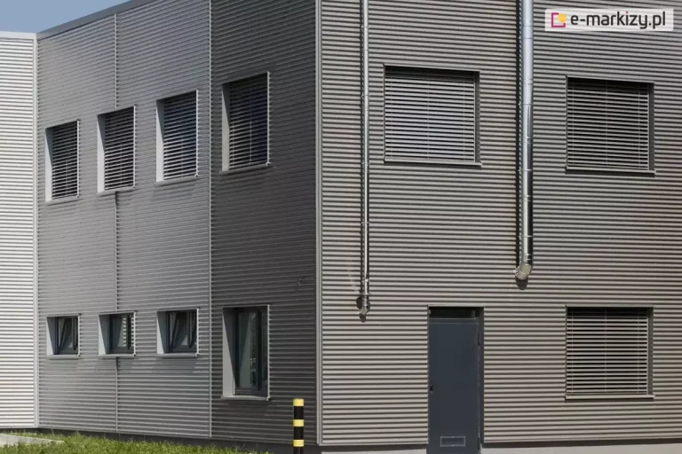 Narożnik nowoczesnego, dwukondygnacyjnego budynku o elewacji z szarej, poziomej blachy trapezowej. Na obu widocznych ścianach zamontowane są zewnętrzne żaluzje fasadowe z poziomymi aluminiowymi lamelami w kolorze srebrnym. Żaluzje osłaniają wszystkie okna na piętrze – cztery od strony lewej oraz trzy od frontu. Dodatkowo, na parterze widoczne są trzy okna bez żaluzji po lewej stronie oraz jedno większe po prawej stronie, także bez osłony. Po lewej stronie narożnika znajduje się słupek ochronny w kolorze żółto-czarnym, ustawiony przed ścianą budynku. Drzwi techniczne w kolorze grafitowym umieszczone są pośrodku prawej ściany. Całość sugeruje obiekt o przeznaczeniu przemysłowym lub biurowym. Podtynkowe żaluzje i ich lamele współgrają z poziomymi pasami elewacji tworząc interesujący efekt.