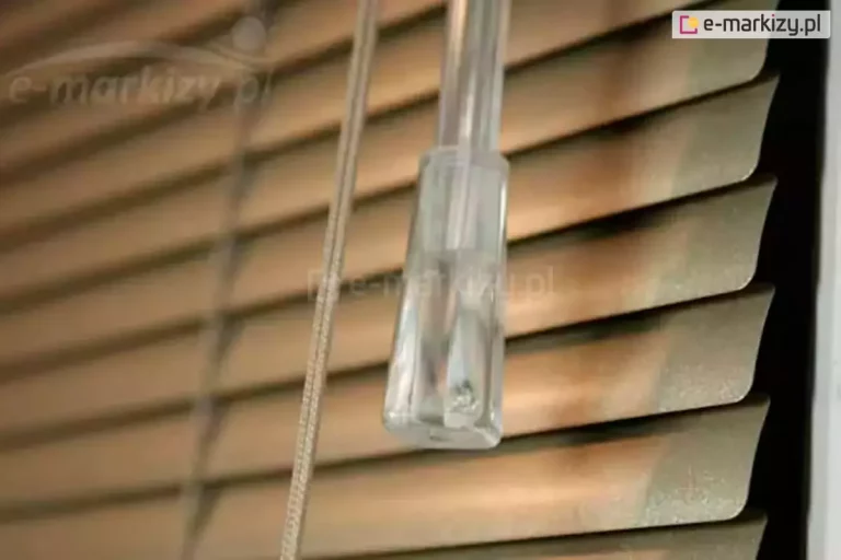 Zbliżenie na detale techniczne sterowania, jakie posiadają żaluzje aluminiowe 16 mm z interesującą teksturą w ciepłym odcieniu brązu. Lamele mają subtelny rysunek wzorów, a ich krawędzie są gładko wykończone i lekko zaokrąglone. Na pierwszym planie widoczny jest pionowy, akrylowy pręt o okrągłym przekroju, służący do precyzyjnego obracania listew i regulacji kąta wpadającego światła. Obok pręta znajdują się białe sznurki operacyjne, które są spięte przezroczystym, plastikowym dzwonkiem o stożkowym kształcie, ułatwiającym chwytanie i podnoszenie całej osłony. W tle widać kolejne poziome segmenty żaluzji ułożone kaskadowo, co tworzy efekt głębi. Oświetlenie pada z lewej strony, akcentując matową powierzchnię aluminium oraz detale mechanizmu sznurkowego. Całość kompozycji skupia się na funkcjonalności i estetyce wykończenia, podkreślając wysoką jakość elementów, z których składa się żaluzja aluminiowa 16 mm.